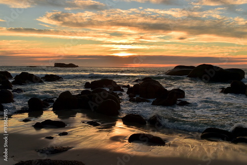 Golden sunset light reflects on the wet sand and granite boulders of Bakoven Beach in Cape Town, South Africa. The Atlantic Ocean waves crash against the rocky coastline under a dramatic sky.