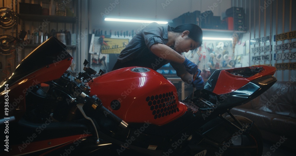 Naklejka premium Professional Female Rider Detaching Battery Cables Using Screwdriver In Workshop. Asian Woman Mechanic in Overalls Works on Electrical Components of Red Motorbike. Concept of Smart Garage Lifestyle.