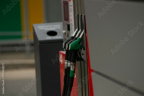 Fuel pump nozzles at urban station green and black hoses resting on metal dispenser beside trash bin, concrete forecourt and payment terminal, city