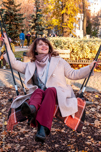 A happy woman of 40-50 years old in a beige coat swings on a swing on an autumn day in the park
