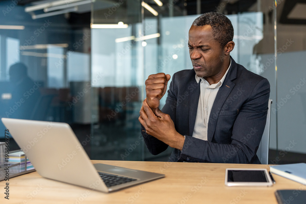 Obraz premium Mature man experiencing discomfort and pain in his wrist while working at his desk with a laptop, indicating potential musculoskeletal problems or carpal tunnel syndrome from prolonged computer use