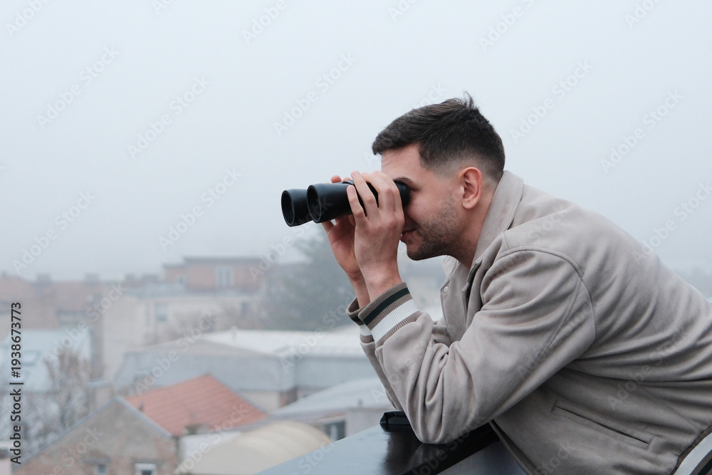 Obraz premium Young man leaning on balcony railing and looking through binoculars over foggy city. Concept of observation, urban exploration and discovering the city view.