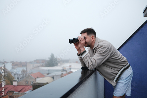 Valokuva Young man leaning on balcony railing and looking through binoculars over foggy city