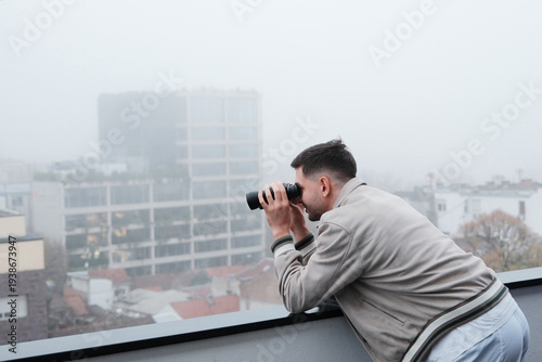 Young man leaning on balcony railing and looking through binoculars over foggy city. Concept of observation, urban exploration and discovering the city view.