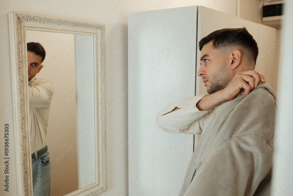 Fototapeta premium Young man looking at his reflection in mirror while adjusting clothes before leaving home. Morning routine and daily lifestyle concept.