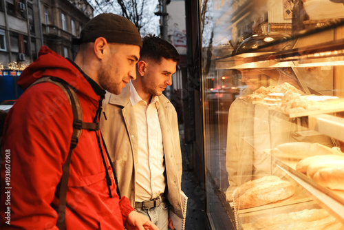 Two men look at fresh bread through bakery window on evening city street. Scene shows everyday urban lifestyle, friendship and food choice during casual city walk.