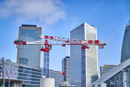 A red and white Potain construction crane stands prominently in the foreground against the modern skyline of La Défense business district in Paris, France. The crane features visible branding