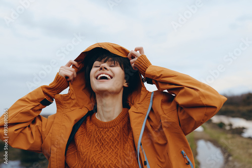 Happy woman lifestyle outdoors wearing orange jacket and sweater adjusts hood in cold weather. Natural background with cloudy sky and blurred landscape. Concept of autumn, joy, and casual fashion.