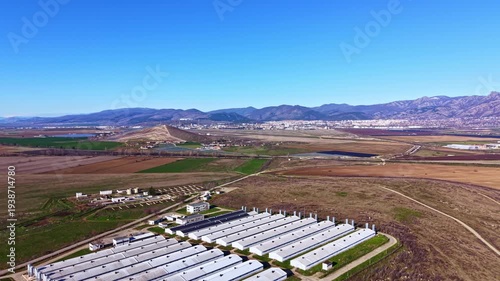 Aerial view shows open fields and buildings in Montana region with mountains in distance under clear blue sky. Land is mostly flat with some agricultural plots and infrastructure visible.