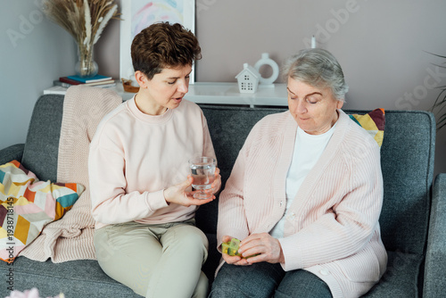Middle aged daughter taking care of old mother, giving pills or vitamin supplements and glass of water helping her mom taking medicine on time. Elderly healthcare and support. Parenting love, care.