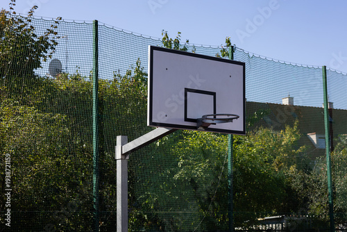 Modern basketball hoop on outdoor sports ground with high safety fence and green trees on background. Professional urban sports infrastructure for active lifestyle and training