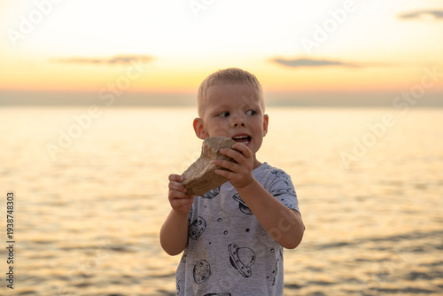 Curious little boy holding big stone rock on beach at golden sunset over sea with excited face expression. Concept of childhood curiosity, outdoor discovery, nature exploration and summer vacation.