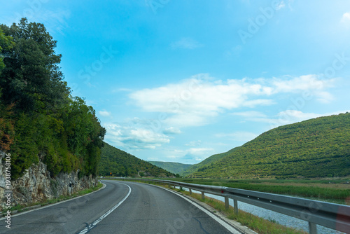 Φωτογραφία Empty curved road alongside rocky cliff and wetland river channel between two green forested hills under bright blue summer sky
