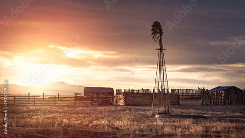 An old, abandoned farm and windmill under a orange evening sky at sunset in the rural countryside.