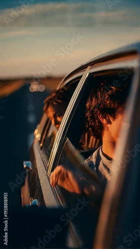 Two smiling young men lean out of car windows into the warm golden light of a sunset road trip, with bokeh highway lights and a broad open landscape glowing in the background.