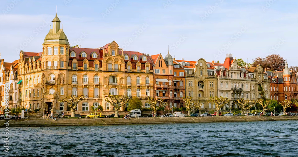 Fototapeta premium Historic buildings line the riverbank in a vibrant city during a sunny afternoon with clear skies and calm waters