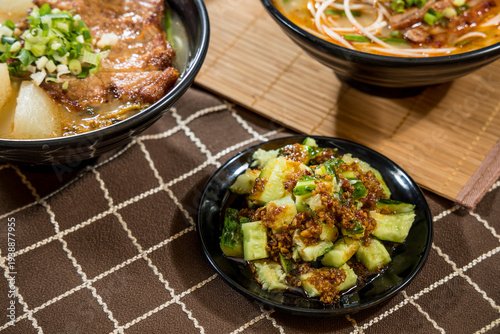 A small black plate holds diced cucumber mixed with garlic, chili, and soy sauce dressing, placed beside a bowl of noodles.