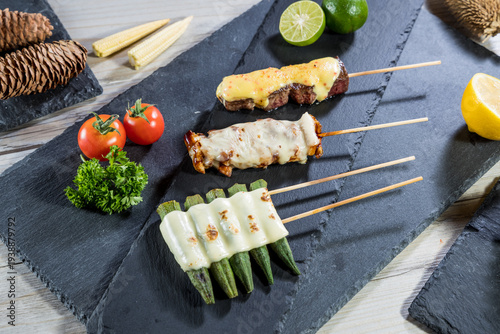 Grilled meat and vegetable skewers topped with melted cheese arranged on a slate board. The creamy texture contrasts with charred flavors ideal for modern barbecue and skewer menu designs.