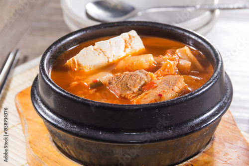 A black stone pot filled with spicy kimchi stew, tofu, and meat served on a wooden board