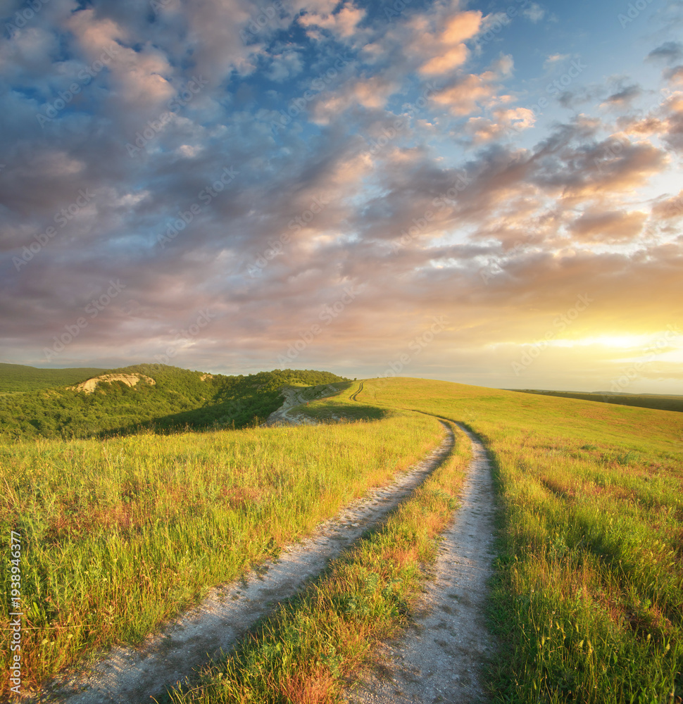Fototapeta premium Rural landscape featuring a winding dirt road leading through lush green hills toward a vibrant sunset sky. A peaceful and atmospheric nature background. Travel, adventure, and freedom-themed designs.