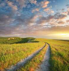 Fotobehang Honingkleur Rural landscape featuring a winding dirt road leading through lush green hills toward a vibrant sunset sky. A peaceful and atmospheric nature background. Travel, adventure, and freedom-themed designs.  © GIS