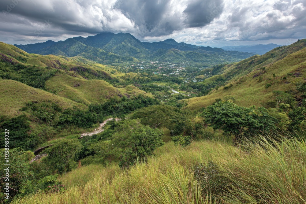 Obraz premium Rolling green hills frame a distant valley town, under dramatic cloudy skies