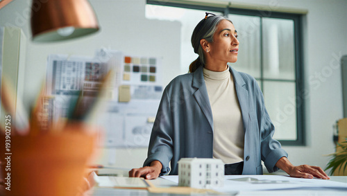 Veteran lead architect taking a contemplative pause from physical desktop models to mentally visualize the real-world scale of her urban project.