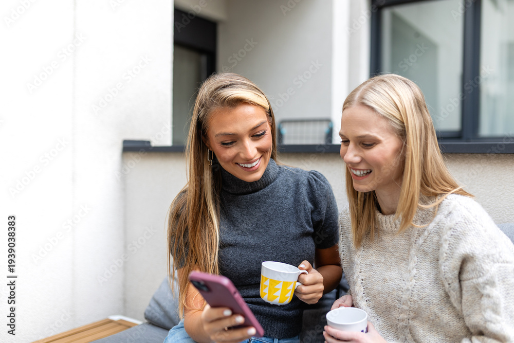 Fototapeta premium Two young female friends using smartphone on balcony. Communication, friendship, and digital lifestyle concept. Happy women looking at phone screen while holding coffee mugs. Social media app.