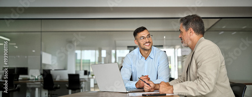 Young smiling professional latin business man working in corporate office with older executive manager using laptop technology having conversation on financial project strategy at work meeting.