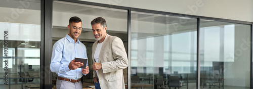 Two happy busy professional business men executive business team working together standing in office holding tab using digital tablet computer tech managing data standing in corporate office.
