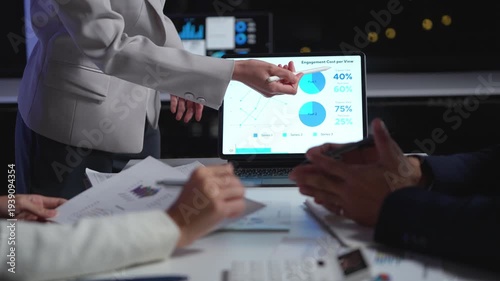 Business people in a modern office meeting, discussing strategy and analyzing charts. A colleague points at a tablet showing graphs and data
