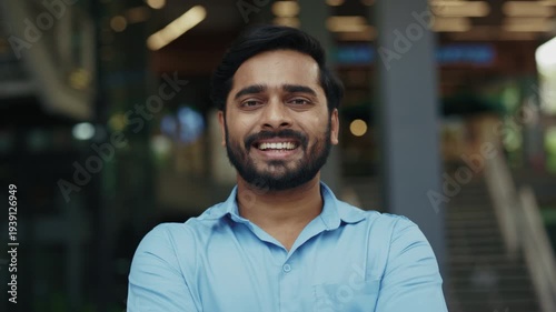 Portrait of positive Indian guy in blue shirt smiles near shopping mall. Young Hindu man in casual attire radiates positive emotions in urban old district. Corporate employee