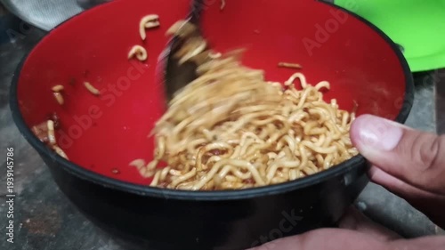 Instant noodles being mixed in a red bowl with a spoon, with a hand holding the bowl.
