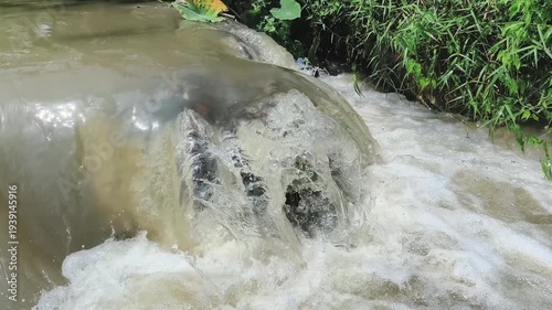 Murky water cascading over rocks, creating a foamy white froth as it flows downstream. 