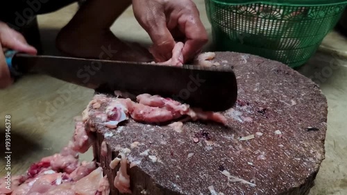 A person's hands using a large knife to chop raw chicken on a wooden cutting board.