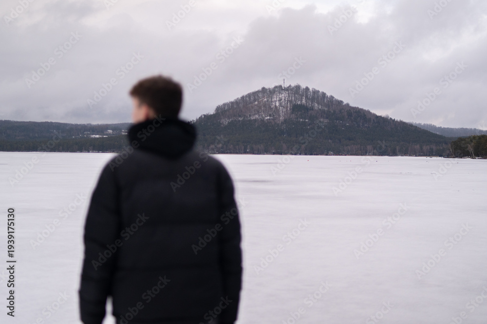 Fototapeta premium View of snowy hill across Macha Lake with blurred man in foreground. Winter landscape in Czech Republic. Man standing near frozen Macha Lake in Czech Republic looking at distant hill. Winter travel an