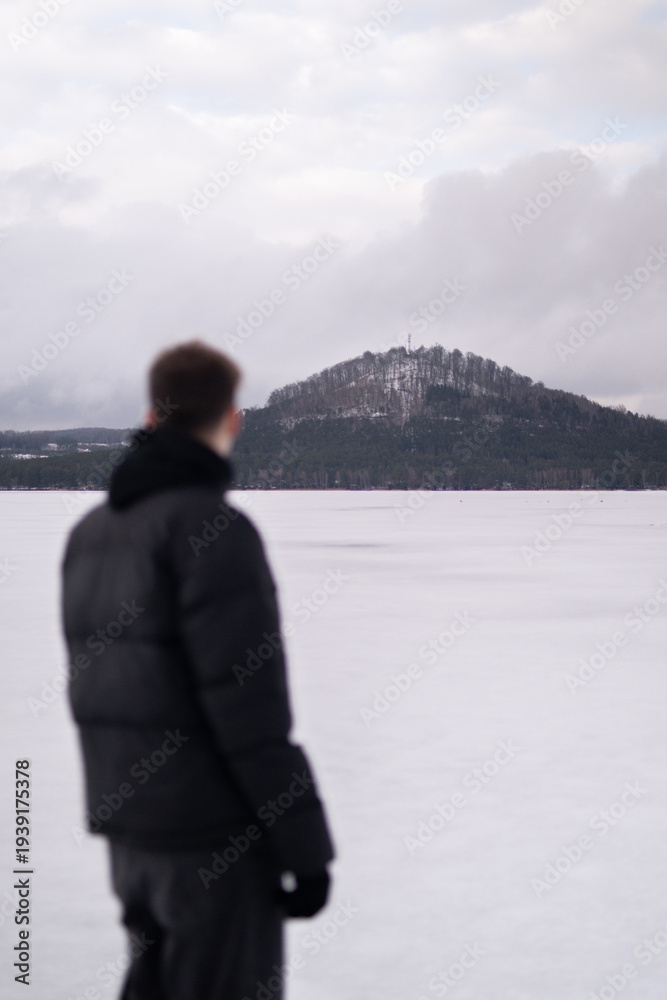 Fototapeta premium View of snowy hill across Macha Lake with blurred man in foreground. Winter landscape in Czech Republic. Man standing near frozen Macha Lake in Czech Republic looking at distant hill. Winter travel an