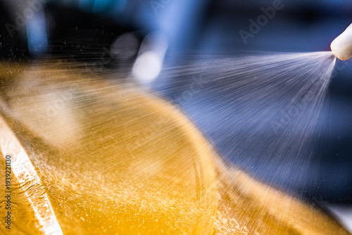 High pressure water spray cleaning yellow car surface in car wash with droplets and reflections visible on the glossy paint finish and blurred background elements