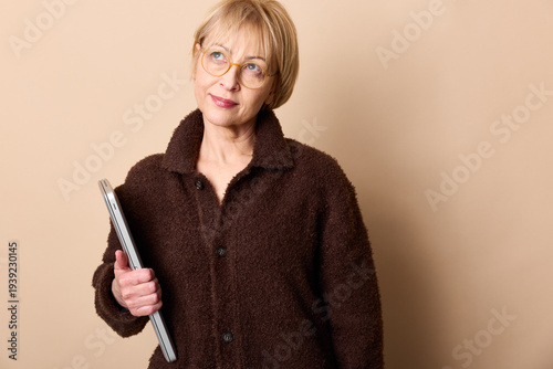 Thoughtful mature woman in glasses holding laptop looks away on beige background. Concept for technology, business, and middle aged professional lifestyle.