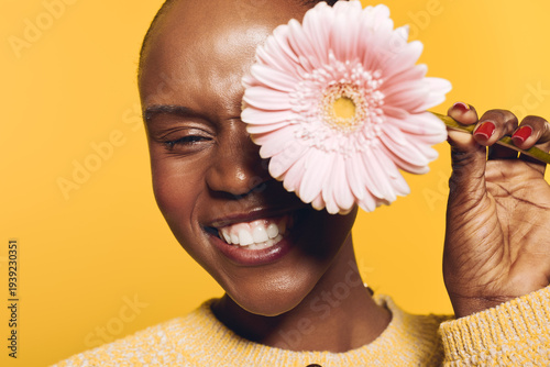 Smiling dark skinned African woman with short hair holds pink flower near face against yellow background. Concept of natural beauty, joy, floral, and positive emotions in studio portrait.