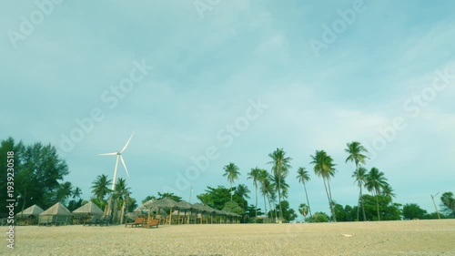 Wind turbine or Windmill on tropical summer sandy beach with palm trees in Pastel cyan blue sky n white  wispy cloud or fluffy cludscape at holiday vacation trip, sustainable n green energy effiency 
