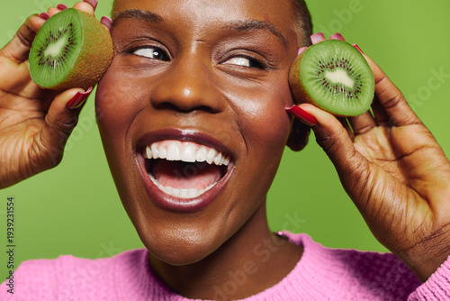 Happy dark skinned African woman with short hair holds two kiwi halves near face smiling broadly on green background. Concept of healthy lifestyle, fresh fruit, and natural beauty.