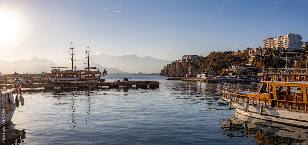Obraz premium Scenic view of Antalya old town harbor Kaleici at bright winter sunset. Coastal landscape with excursion boats, mediterranean sea, rocky cliffs and mountains in haze. Antalya, Turkey.