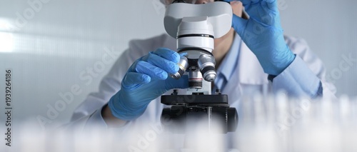 Quadro su tela Woman scientist wearing lab coat, white protective gloves and glasses, is using microscope in modern laboratory setup, portrait view