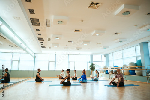 People meditating, sitting in Lotus pose, doing yoga exercises together with trainer in gym
