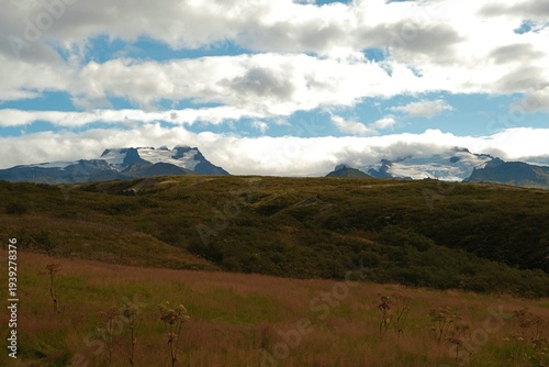 Fotografie Wide Icelandic landscape with grassy hills and distant glaciers of Vatnajokull under dramatic clouds