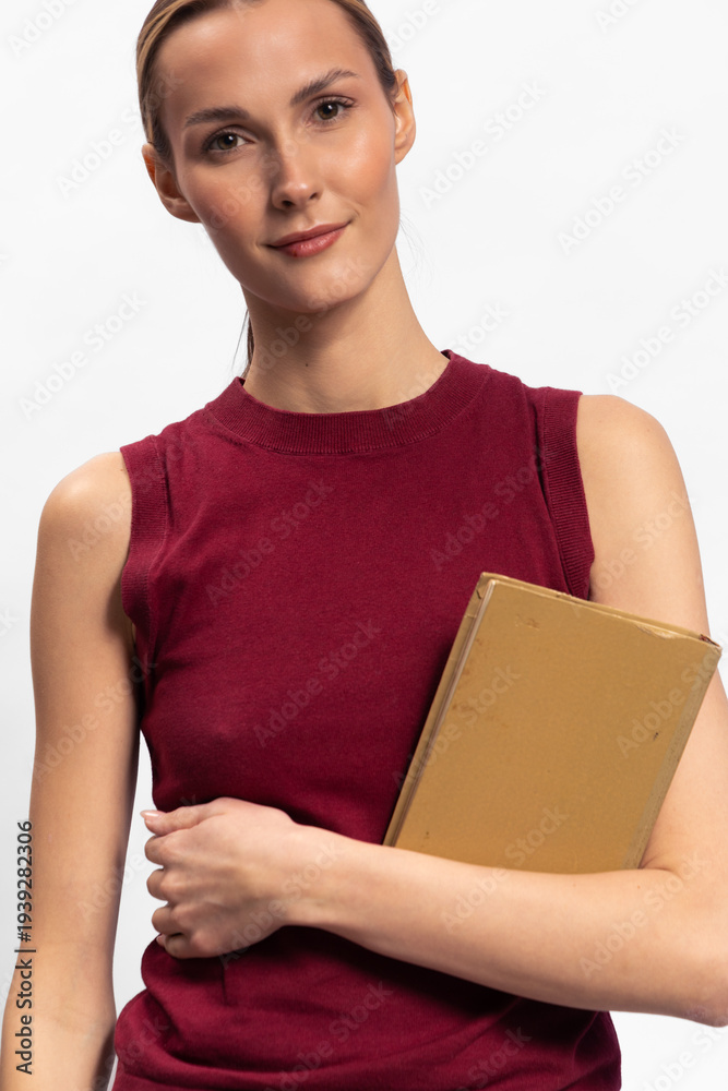 Obraz premium Young woman in sleeveless burgundy top holds closed book and looks at camera with slight smile. Isolated on white background. Concept for education, knowledge, and confidence.