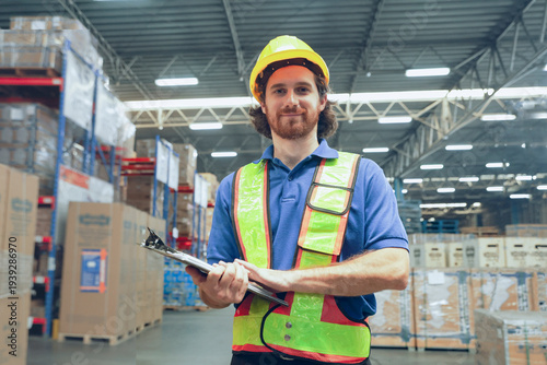 Man wearing helmet and reflective vest is easily noticeable while inspecting warehouse in a Southeast Asian logistics system. He stands cleverly holding clipboard preparing goods for shipment.