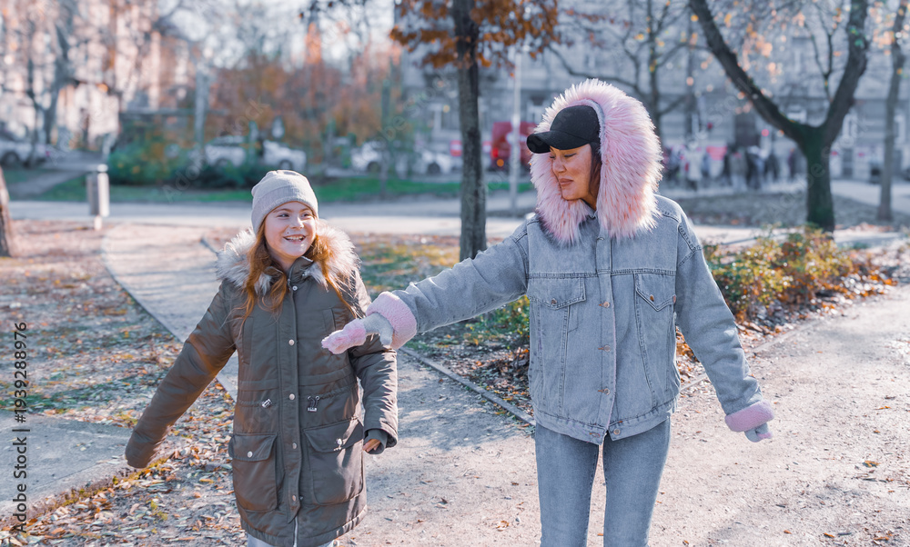 Obraz premium Happy mother and ten year old daughter smiling and playing together in sunny autumn urban park while holding hands with soft bokeh background and warm lens flare on bright day with copy space
