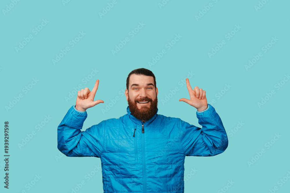 Fototapeta premium Playful Caucasian Man Pointing Upward With Smile, Bright Turquoise Background And Blue Jacket Conveying